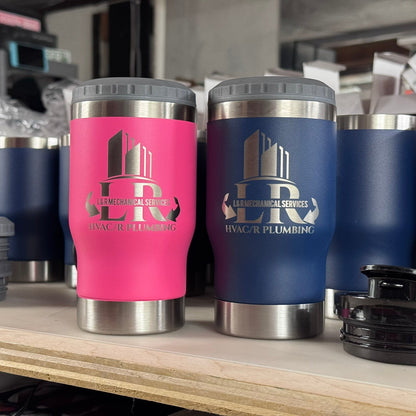 Two insulated stainless steel travel tumblers with gray lids sit on a wooden shelf in a workshop. The left tumbler is hot pink with a silver logo reading 'LR Le Mechanical Services HVAC/R Plumbing', and the right tumbler is dark blue with a matching silver logo.